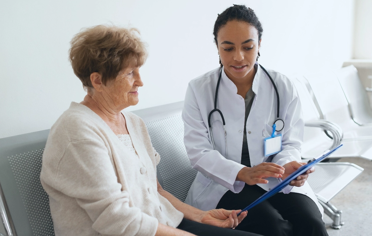 Female healthcare professional and female patient sitting on a sofa looking at a paper on a clipboard.